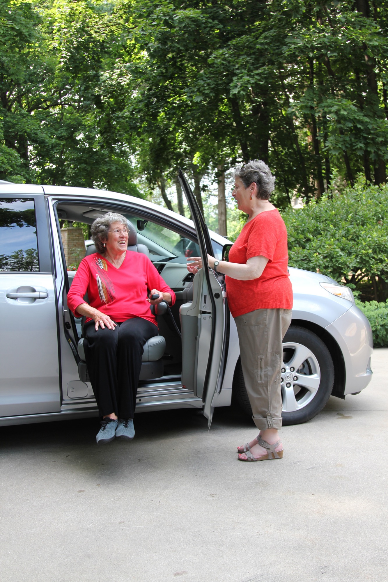 A seated older woman on a Turny Evo transfer seat extended outside a silver Toyota Sienna in a suburban driveway; a companion stands beside her holding the door.